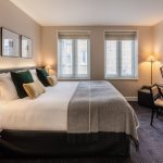 A modern hotel room with a large double bed, neatly made with white linens and decorative pillows. The room features two windows, wall art, a flat-screen TV, a desk with a lamp, and warm ambient lighting from floor lamps.