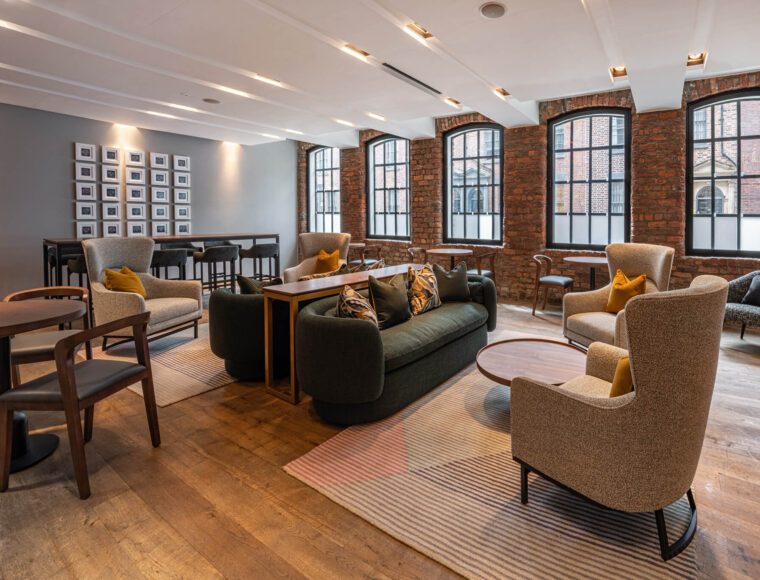 A modern lounge area at The Resident's Liverpool City Centre hotel, features stylish furniture against a backdrop of brick walls with arched windows. The room includes sofas, armchairs, and coffee tables, with a gallery wall of photos and a high table with stools. The wooden floor is complemented by cozy area rugs.