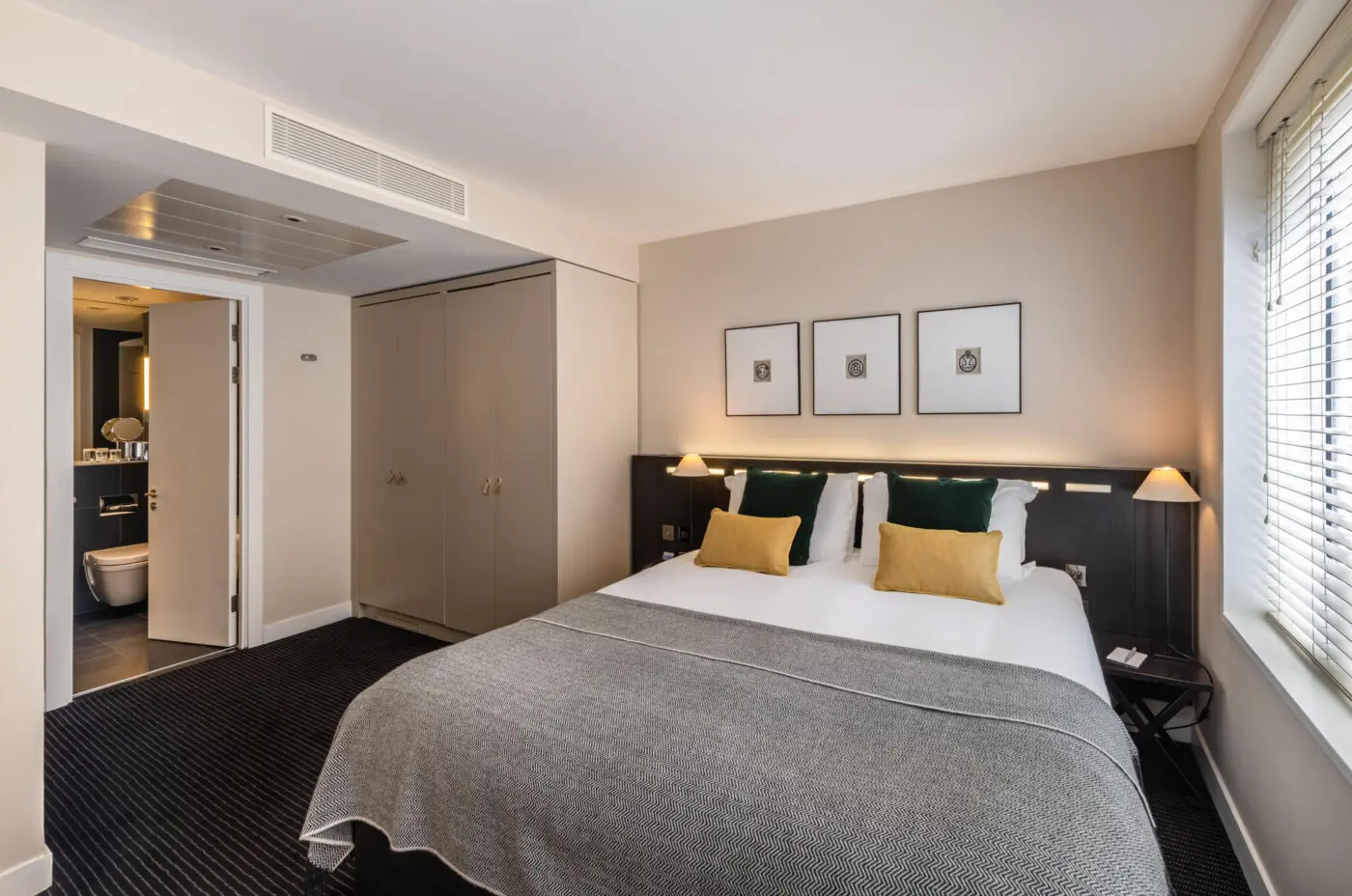 A modern bedroom at The Resident Soho features a large bed with white linens, mustard and green pillows, and a grey throw. Two bedside tables with lamps flank the bed. The room includes a large closet, a partially open door leading to a bathroom, and three framed artworks above the bed.