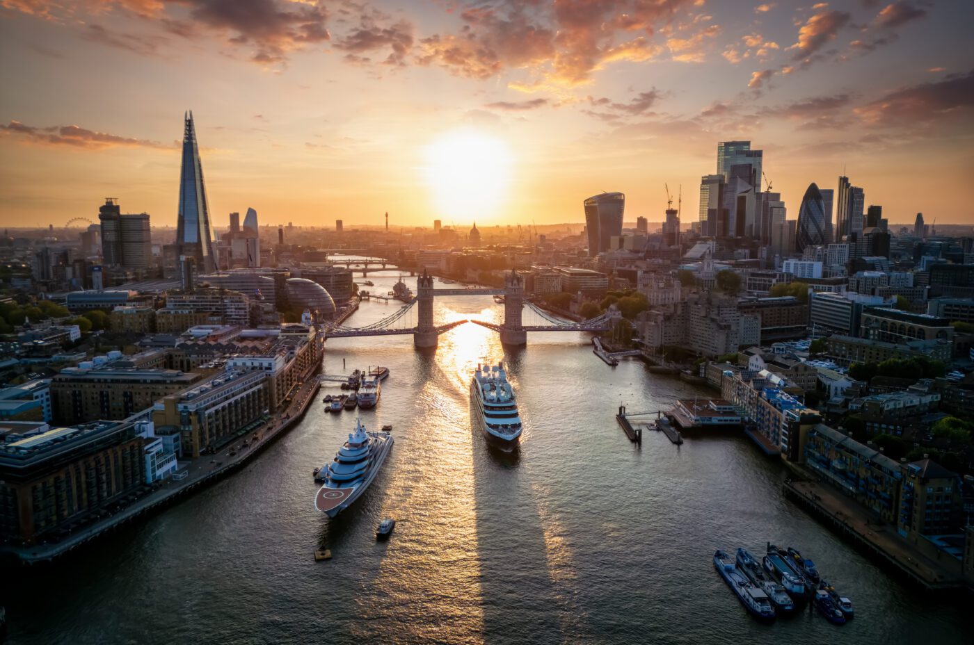 An aerial view of London at sunset, featuring the Thames River, Tower Bridge, and the city's skyline with notable buildings like The Shard. Boats for sale are visible on the river, and the sky is partly cloudy with warm sunlight casting long shadows.