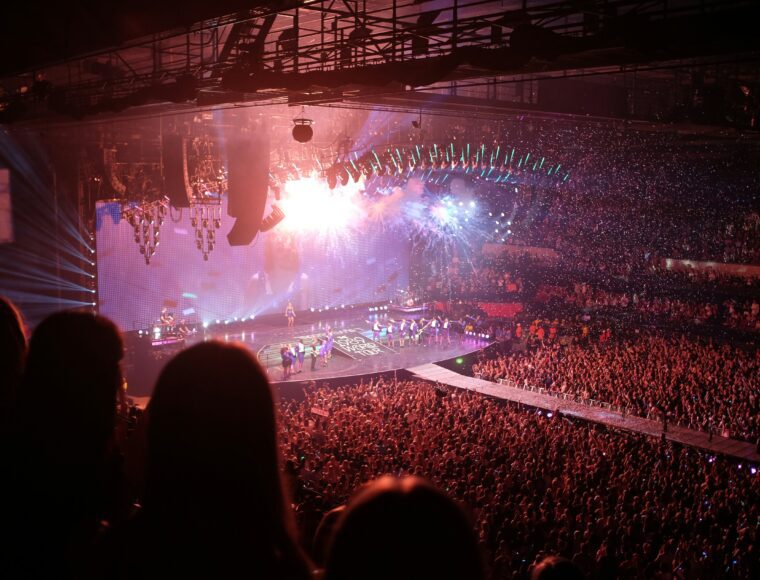 A large, energetic crowd is at an indoor concert showing Taylor Swift Bright lights illuminate the stage where performers are actively engaging the audience. The venue is packed with spectators, and the atmosphere is lively with various lights and effects enhancing the show.