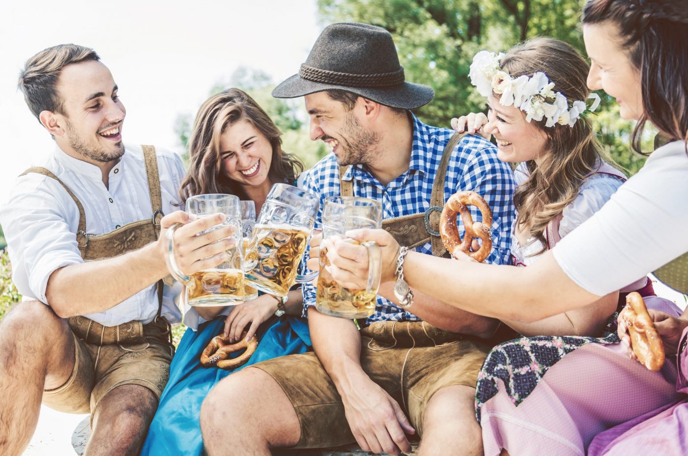 Four people dressed in traditional Bavarian clothing are sitting outdoors, cheerfully toasting with large beer steins during Oktoberfest.