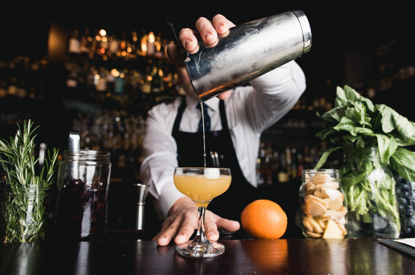 A bartender in a white shirt and black apron pours a cocktail from a shaker into a glass at the bustling Fringe Festival. The bar counter is adorned with jars of garnishes, a fresh orange, and herbs.