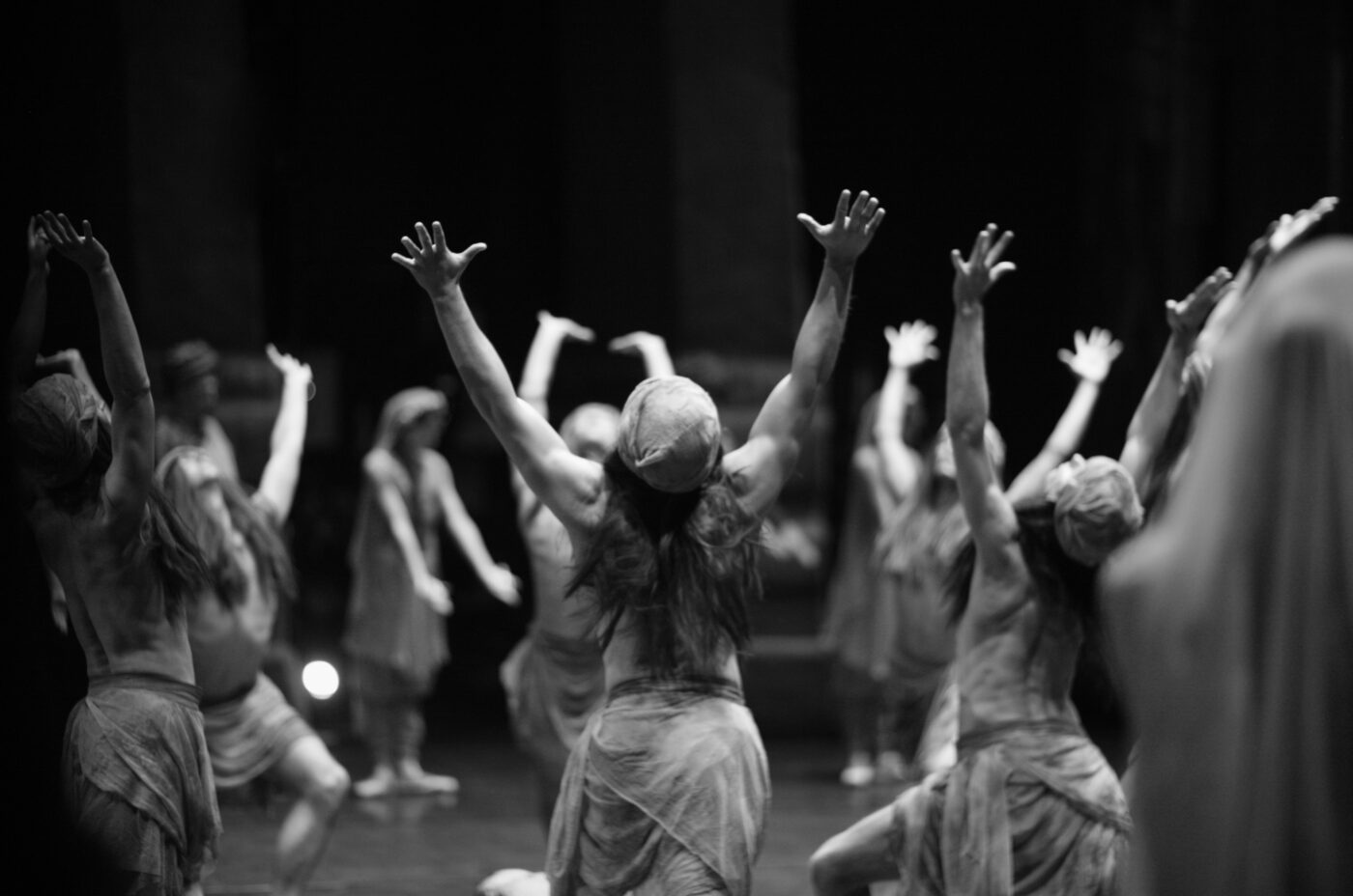 A black and white photo captures a group of dancers performing on stage at the Storytelling Festival. The dancers, wearing flowing costumes and headscarves, have their arms extended upward. The scene is dramatic, with lighting highlighting their expressive movements.