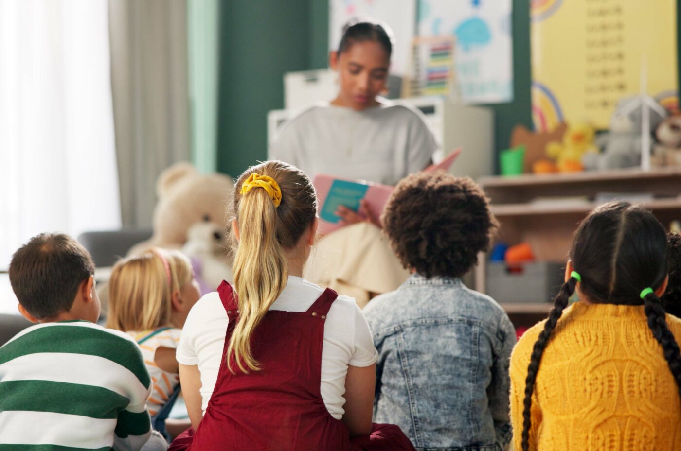 A woman is reading a book to a group of young children sitting on the floor. The children, with various hairstyles and colorful clothing, are attentively facing her. The room is colorful, reminiscent of The Edinburgh Festival, with children's artwork and educational posters on the walls.