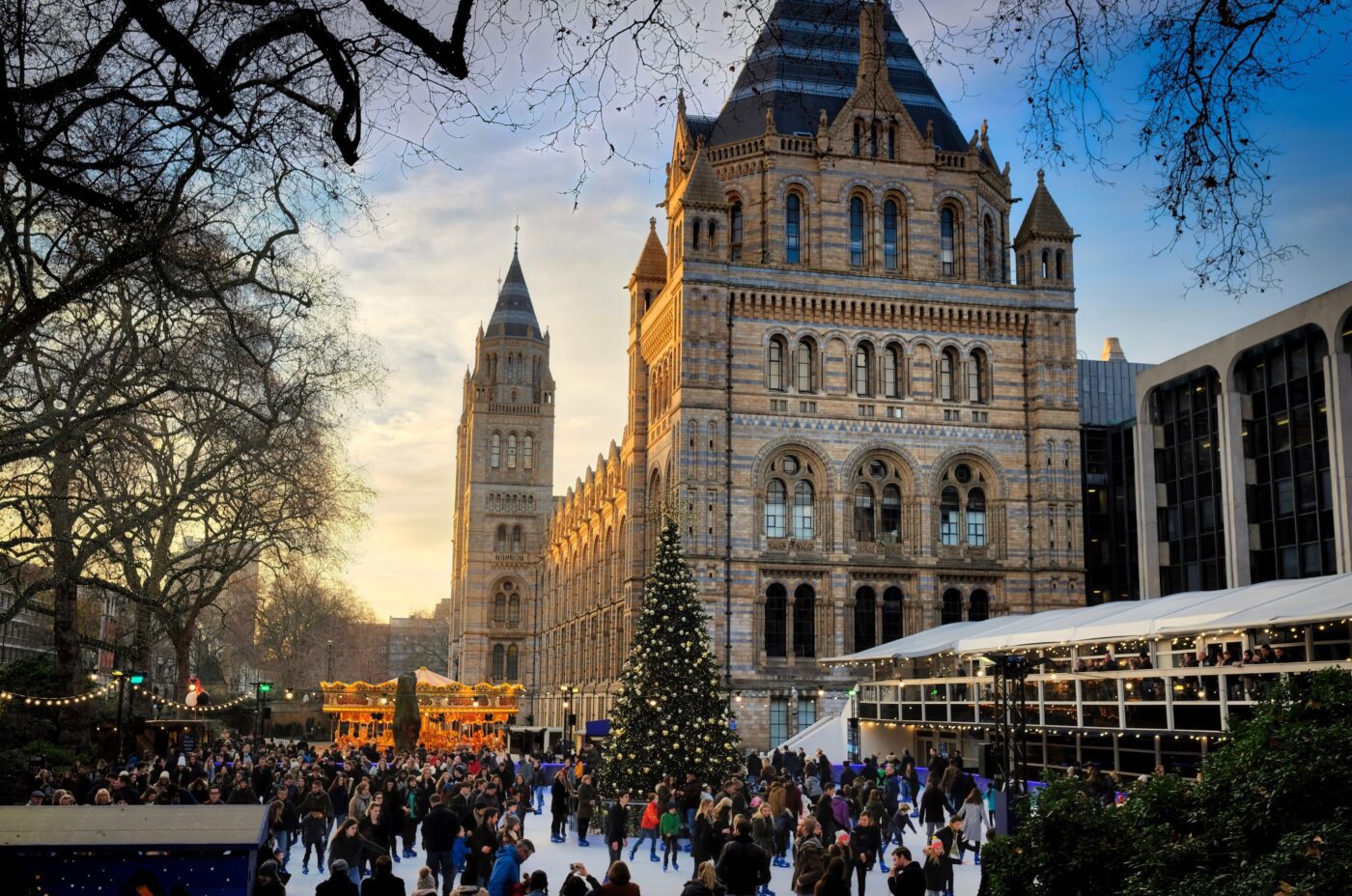 A festive scene with a large decorated Christmas tree and a bustling ice skating rink set against the backdrop of a historic building. People are enjoying the winter atmosphere, and a carousel is visible nearby. Bare trees frame the scene under a blue sky.