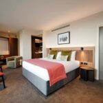 A modern hotel room with a large bed, red blanket, and green cushions. The room features a seating area with two chairs and a small table. A closet and a bathroom are visible in the background, with neutral-colored walls and soft lighting.