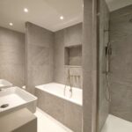 A modern bathroom featuring a gray tiled interior with a white bathtub, a glass shower area, and two white sinks with silver faucets. The space is well-lit with recessed ceiling lights.