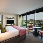 A modern hotel room with a king-sized bed adorned with white sheets and pink accents. A wall-mounted TV faces the bed. Large windows showcase a cityscape view with buildings under a cloudy sky. Two chairs and a small round table sit nearby.