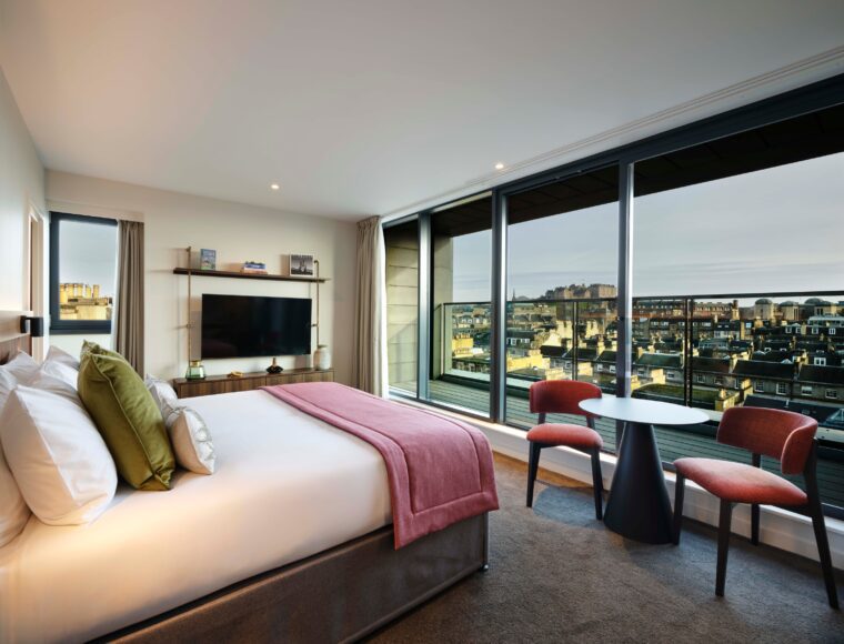 A modern hotel room with a king-sized bed adorned with white sheets and pink accents. A wall-mounted TV faces the bed. Large windows showcase a cityscape view with buildings under a cloudy sky. Two chairs and a small round table sit nearby.