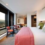 Hotel room with floor-to-ceiling windows leading to a balcony. The room features a bed with red and green accents, a seating area with chairs and a table, and modern decor. Cityscape views are visible through the windows.