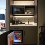 A compact kitchenette with a microwave, shelves with glassware, a sink, coffee maker, kettle, and a small fridge stocked with drinks and fruit. An umbrella stand is visible on the right.