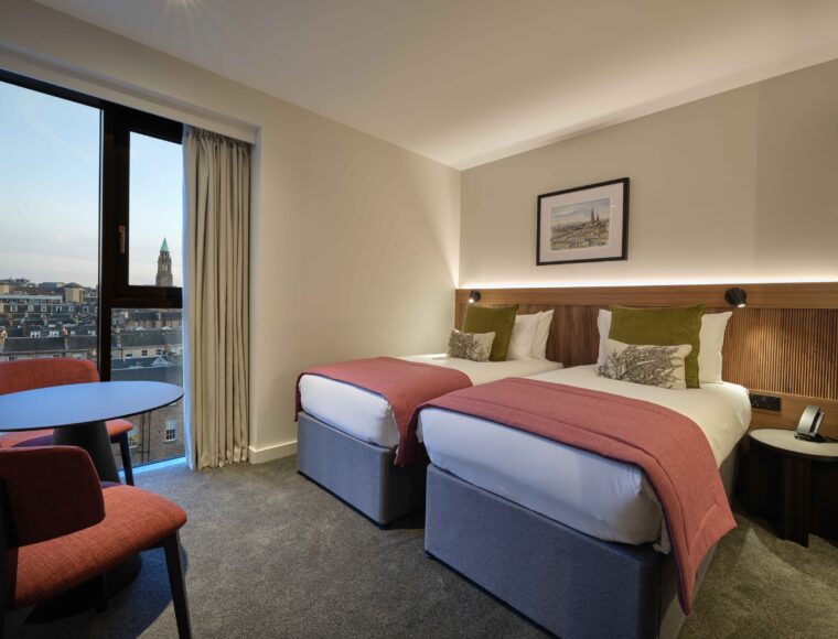 A hotel room with two neatly made twin beds topped with red throws and green pillows. A framed picture hangs above the headboards. A small table with two chairs is by the window, which offers a view of city rooftops.