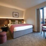 A cozy hotel room with a large bed adorned with white and red bedding. The room features a wooden headboard, a wall lamp, a circular mirror, and a round table with two chairs by a window offering a view of a cityscape.