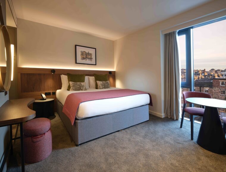 A cozy hotel room with a large bed adorned with white and red bedding. The room features a wooden headboard, a wall lamp, a circular mirror, and a round table with two chairs by a window offering a view of a cityscape.