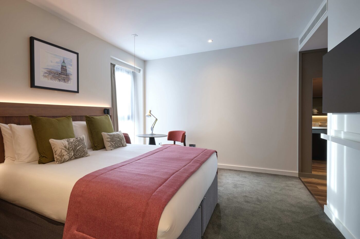 A modern hotel room featuring a large bed with white linens, green and white pillows, and a red blanket. A wooden headboard adorns the bed. There's a desk with a lamp, a chair, and a wall-mounted art piece. The room has soft lighting and carpeted flooring.