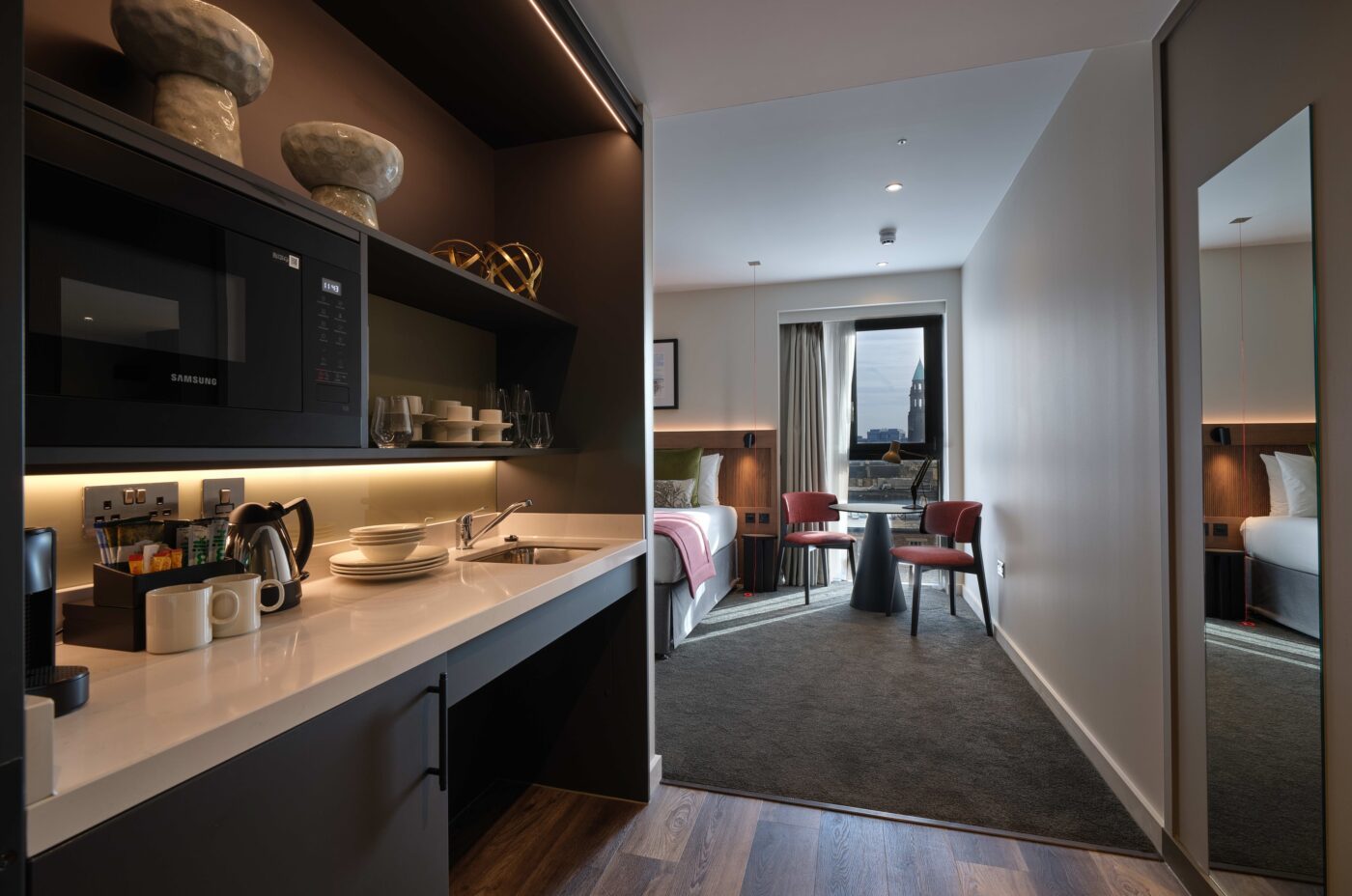 A modern hotel suite with a kitchenette on the left featuring a microwave, sink, and neatly arranged dishes. In the background, a cozy bedroom with a neatly made bed, armchairs, a table, and a large window with a city view.