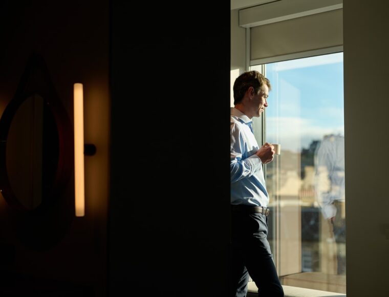 A man standing in a sunlit room looks out a window while holding a cup. He is partially obscured by a dark wall, creating a contrast between light and shadow. The outdoor scene through the window is blurred.