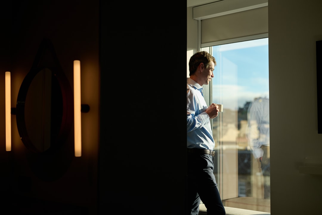 A man standing in a sunlit room looks out a window while holding a cup. He is partially obscured by a dark wall, creating a contrast between light and shadow. The outdoor scene through the window is blurred.