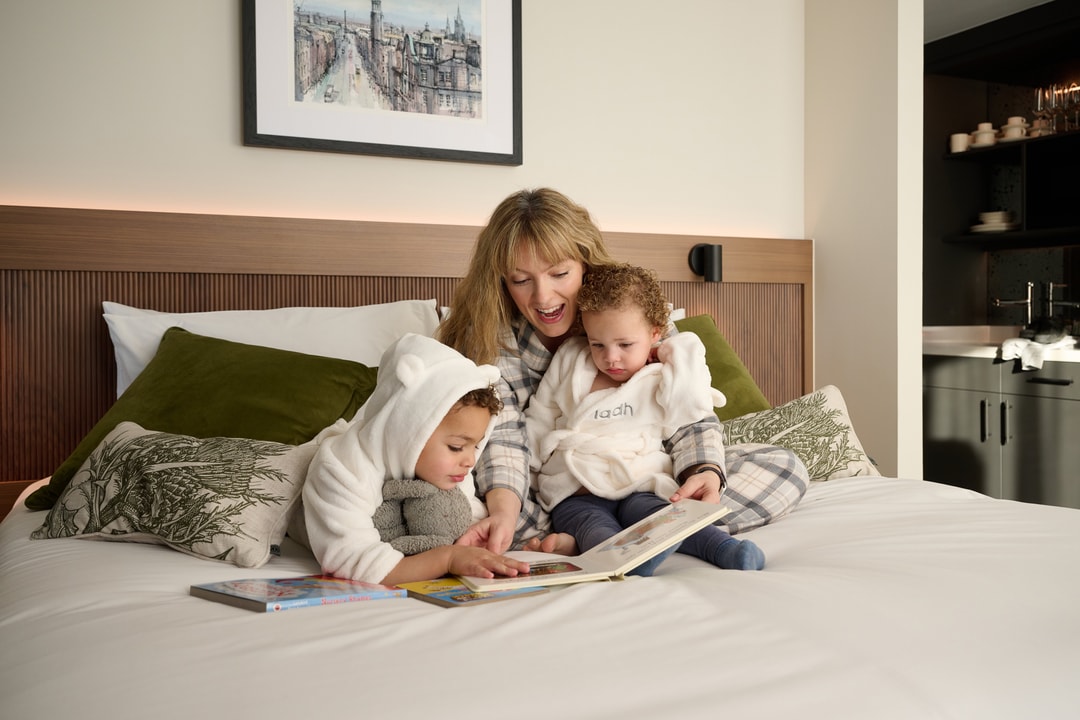 A woman and two small children sit on a bed. The woman is reading a book to the children, who are wearing cozy, hooded outfits. The bed is adorned with green pillows, and a framed picture hangs on the wall behind them.