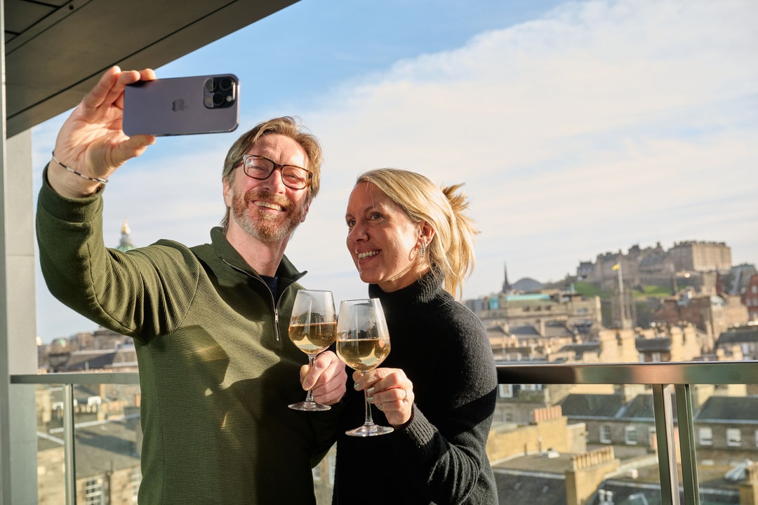 A man and woman stand on a balcony overlooking a cityscape, smiling and taking a selfie. They are each holding a glass of white wine. The background shows buildings and a clear sky.