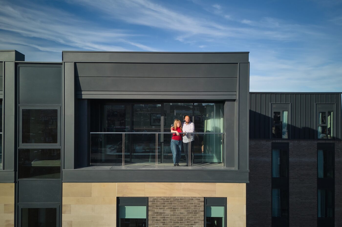 A couple stands on the balcony of a modern building under a clear blue sky. They are looking out from a glass railing, enjoying the view. The structure has a contemporary design with large windows and a mix of dark panels and brickwork.