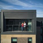 A couple stands on the balcony of a modern building under a clear blue sky. They are looking out from a glass railing, enjoying the view. The structure has a contemporary design with large windows and a mix of dark panels and brickwork.