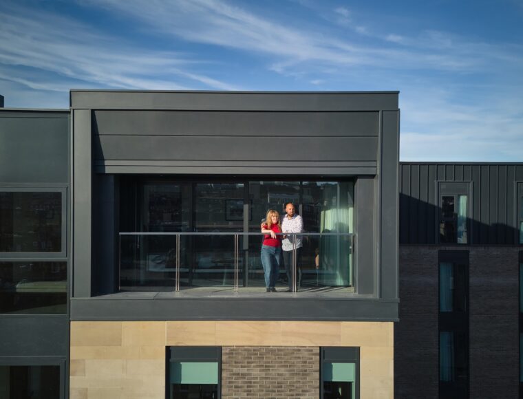 A couple stands on the balcony of a modern building under a clear blue sky. They are looking out from a glass railing, enjoying the view. The structure has a contemporary design with large windows and a mix of dark panels and brickwork.