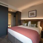 Modern hotel room with a large bed featuring white linens, green and patterned pillows, and a pink throw blanket. A framed artwork hangs on the light beige wall. Dark wood accents and soft lighting create a cozy atmosphere.