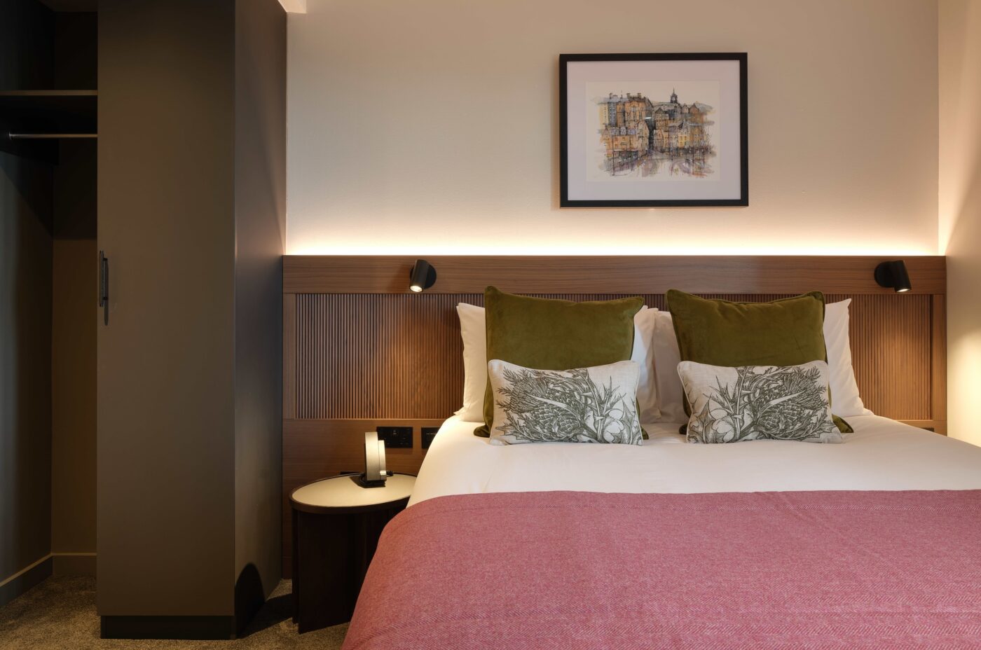 A cozy hotel room with a neatly made double bed covered in white and pink linens, green pillows, and decorative cushions. A framed artwork hangs above the bed. A small round bedside table with a lamp is on the side. The room has warm ambient lighting.