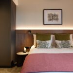 A cozy hotel room with a neatly made double bed covered in white and pink linens, green pillows, and decorative cushions. A framed artwork hangs above the bed. A small round bedside table with a lamp is on the side. The room has warm ambient lighting.