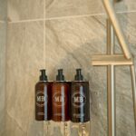 Three wall-mounted shower dispensers containing toiletries are installed against gray stone tiles. The dispensers have black bottles with pumps, labeled 