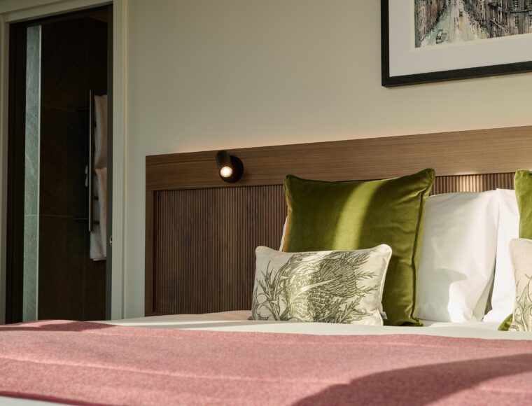 A cozy bedroom features a neatly made bed with green and patterned pillows, a pink blanket, and a wooden headboard. A framed picture hangs on the wall, and sunlight streams in from a nearby window, creating a warm atmosphere.