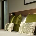 A cozy bedroom scene featuring a bed with two large green velvet pillows and two smaller decorative pillows with leaf patterns. Soft sunlight filters through a window, casting warm light on the bed's wooden headboard and white sheets.