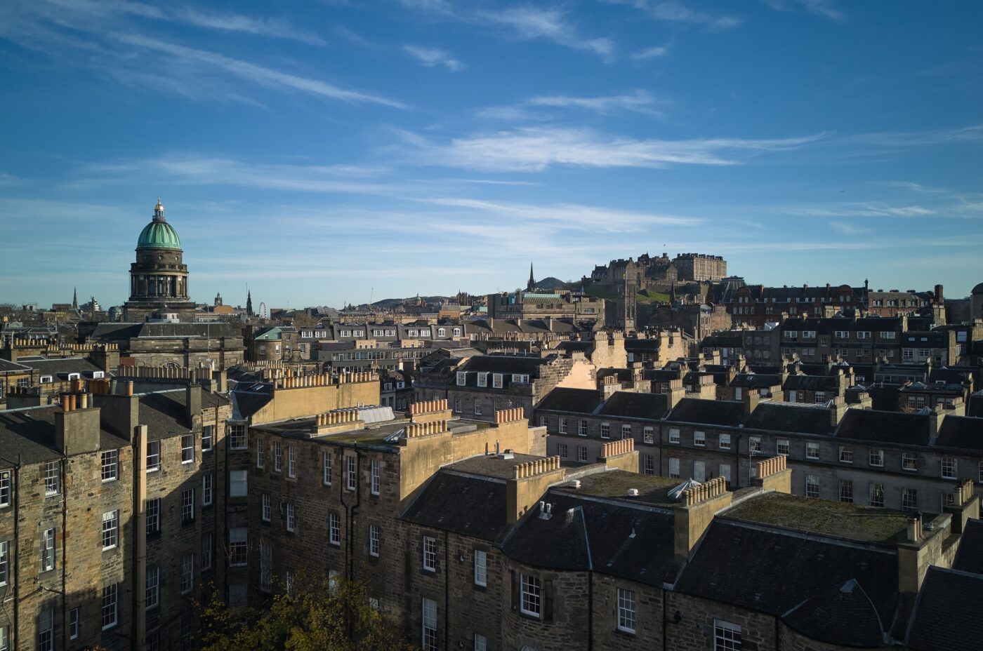 Aerial view of a historic cityscape with stone buildings under a blue sky. A prominent dome and an ancient castle are visible in the background, surrounded by rooftops.