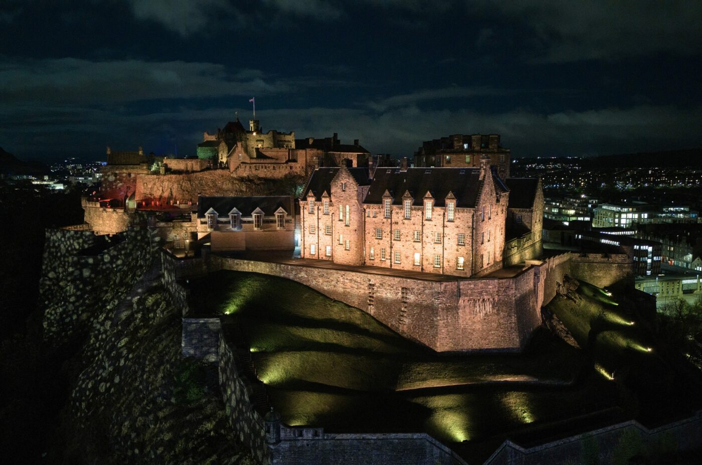 Aerial view of an illuminated castle at night, situated on a rugged hill. The cityscape below is dotted with lights, and the night sky is partially clouded, enhancing the dramatic atmosphere.