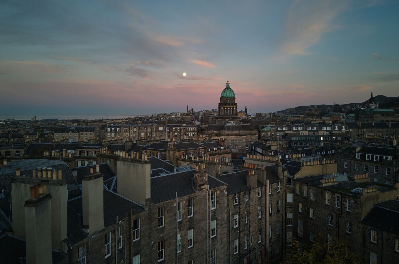 A panoramic view of Edinburgh at dusk, showcasing historic stone buildings with chimneys, a prominently domed structure, and hills in the background under a pink and blue sky with the moon visible.