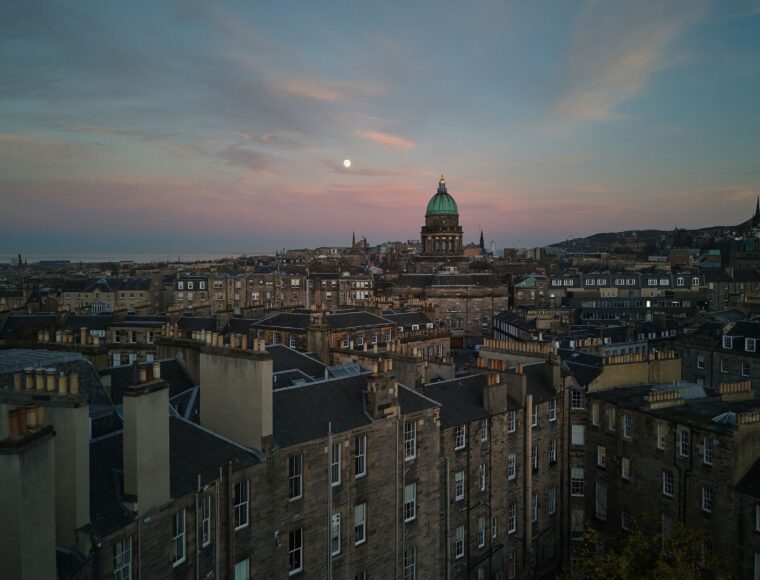 A panoramic view of Edinburgh at dusk, showcasing historic stone buildings with chimneys, a prominently domed structure, and hills in the background under a pink and blue sky with the moon visible.