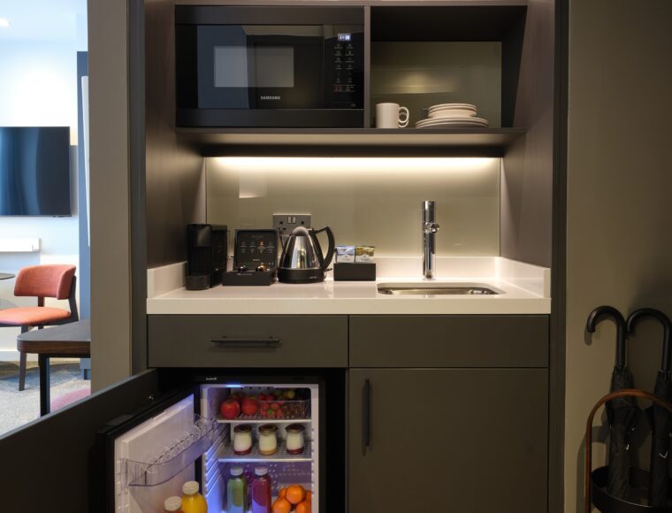 Compact kitchenette with dark cabinets, open mini fridge containing drinks and fruits, microwave, and shelving with glasses and plates. A kettle and coffee machine sit on the counter. An umbrella is propped against the wall.