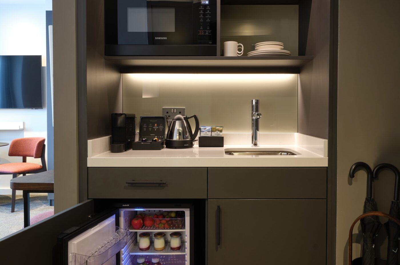 Compact kitchenette with a microwave, coffee maker, kettle, and sink set in dark cabinetry. An open mini-fridge below reveals drinks and fruits. Shelves hold glasses and plates. An umbrella is propped up nearby.