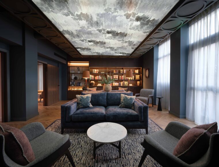 A modern living room with a dark, moody ambiance features a blue sofa, two gray armchairs, and a round marble coffee table on a patterned rug. The ceiling is artistically decorated. A wall lined with bookshelves and decorative items is in the background.