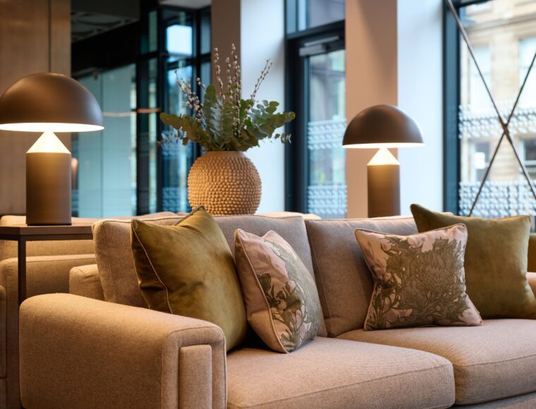 A modern living room with a beige sofa adorned with green and floral cushions. Two matching lamps with dome-shaped shades stand on either side. A large woven vase with leafy plants decorates the background. Large windows allow natural light in.