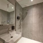 A modern bathroom featuring a large mirror, a white sink with a silver faucet, and a walk-in shower with a glass partition. The shower has a rainfall showerhead and tiled walls in a gray stone pattern.