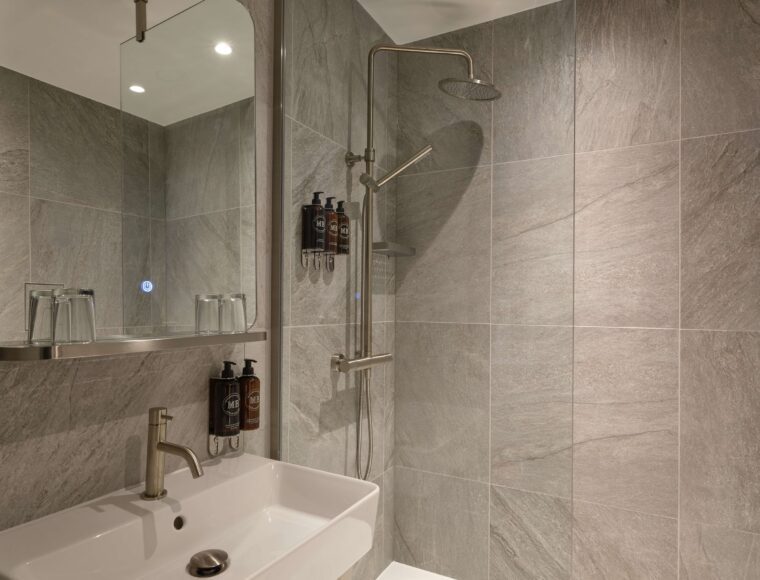 A modern bathroom featuring a large mirror, a white sink with a silver faucet, and a walk-in shower with a glass partition. The shower has a rainfall showerhead and tiled walls in a gray stone pattern.