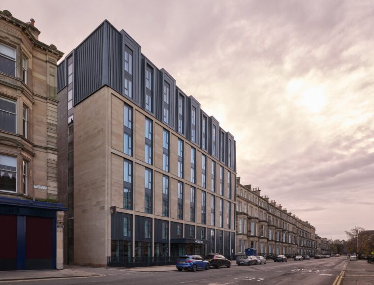 A street view of a modern, multi-story building with large windows, this architectural marvel harmonizes stone and metal cladding. Nestled alongside traditional stone structures under a partly cloudy sky, it stands proudly near the vibrant venues of the Edinburgh International Festival.