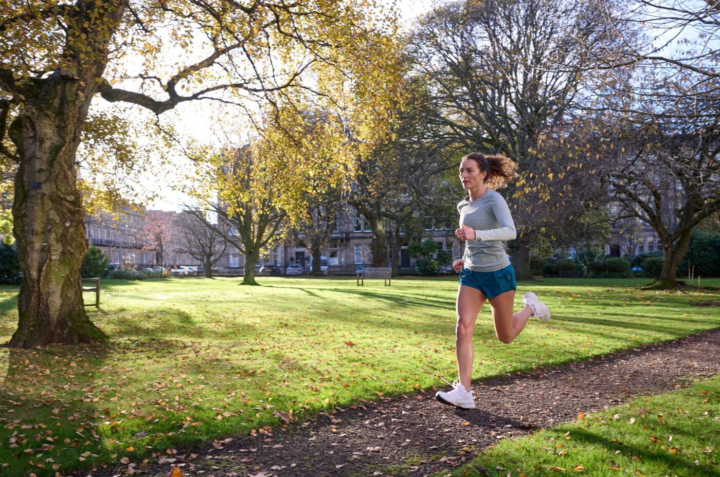 A woman is jogging on a path in a park, surrounded by trees with autumn foliage under a bright, clear sky. She wears a light blue long-sleeve shirt, teal shorts, and white sneakers. The air carries a hint of festive anticipation as the nearby Christmas market begins to stir with life.