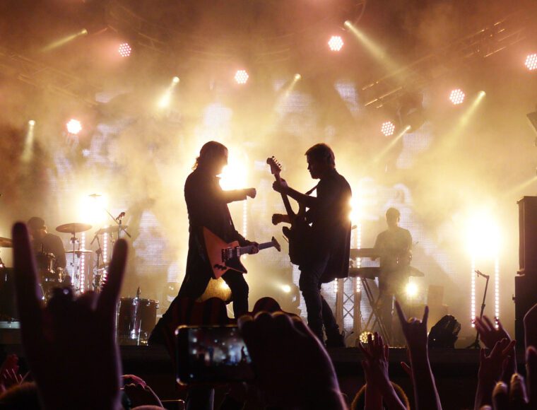 Silhouettes of two guitarists performing on a brightly lit stage during International Beatles Week, surrounded by fog and lights. A crowd in the foreground raises hands and phones, capturing the moment. Drummer and keyboardist are visible in the background.