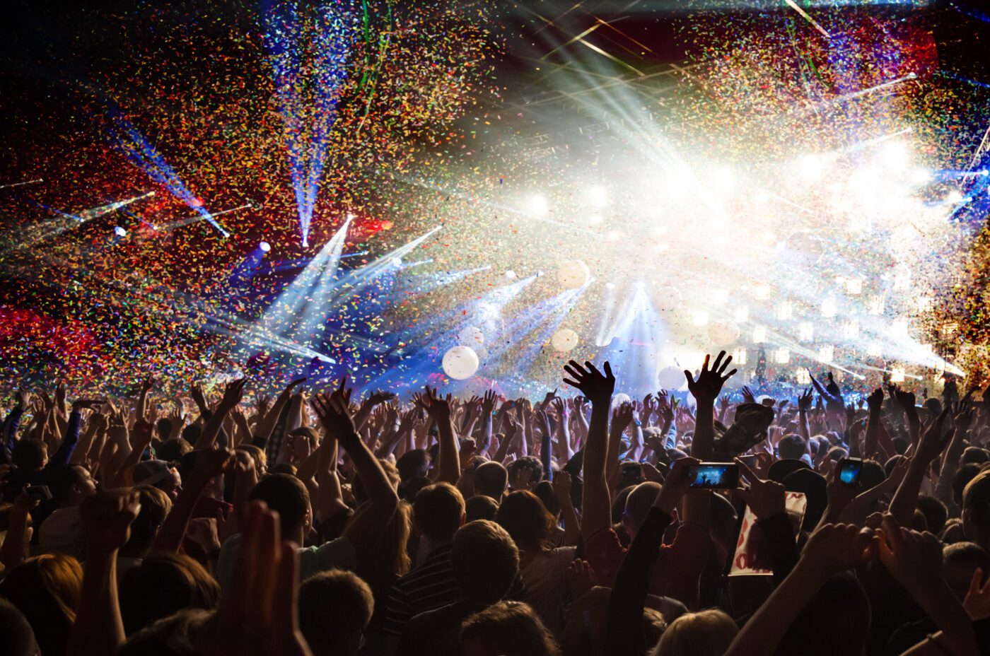 A vibrant concert scene during International Beatles Week features a crowd raising their hands under colorful confetti. Bright stage lights illuminate the area, creating an energetic atmosphere as fans capture the moment with their phones.